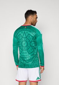 Πράσινη μακρυμάνικη φανέλα ποδοσφαίρου με κυκλικά σχέδια και υφή. Διαθέτει κόκκινες και λευκές λεπτομέρειες στον γιακά και στα μανίκια. Λευκό σορτσάκι.