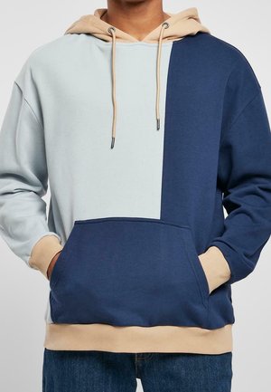 Homme portant un sweat à capuche color-block avec capuche beige, côté gauche bleu clair, côté droit bleu foncé, poche avant, poignets et ourlet beiges.