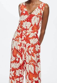 Červený květinový overal s výstřihem do V, bílými botanickými vzory, páskem v pase a kapsami na obou stranách. Vyrobeno z lehké látky.