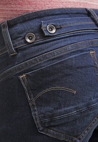 Jeans de mezclilla azul oscuro con dos botones metálicos en la cintura, bolsillos traseros cosidos y costuras visibles.