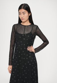 Čierne dlhé šaty s dlhým rukávom z priehľadnej látky s vystúpenými čiernymi bodkami, so zaobleným výstrihom a priliehavým živôtikom.