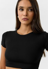 Zwarte crop top met korte mouwen, gemaakt van een gladde stof. De top heeft een ronde hals en een aansluitend ontwerp, met zichtbare stikdetails langs de zoom.