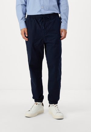 Pantalones azul marino, con cintura elástica y cordón, con puños ajustados y bolsillos laterales, hechos de una tela ligera. Llevados con zapatillas blancas.