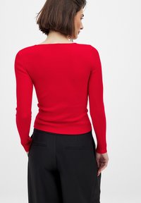 Punainen ribattu pitkähihainen paita, jossa on neliömäinen kaula-aukko, yhdistetty mustiin korkeavyötäröisiin housuihin, joissa on huomaamattomat taskut. Silkkinen pinta.