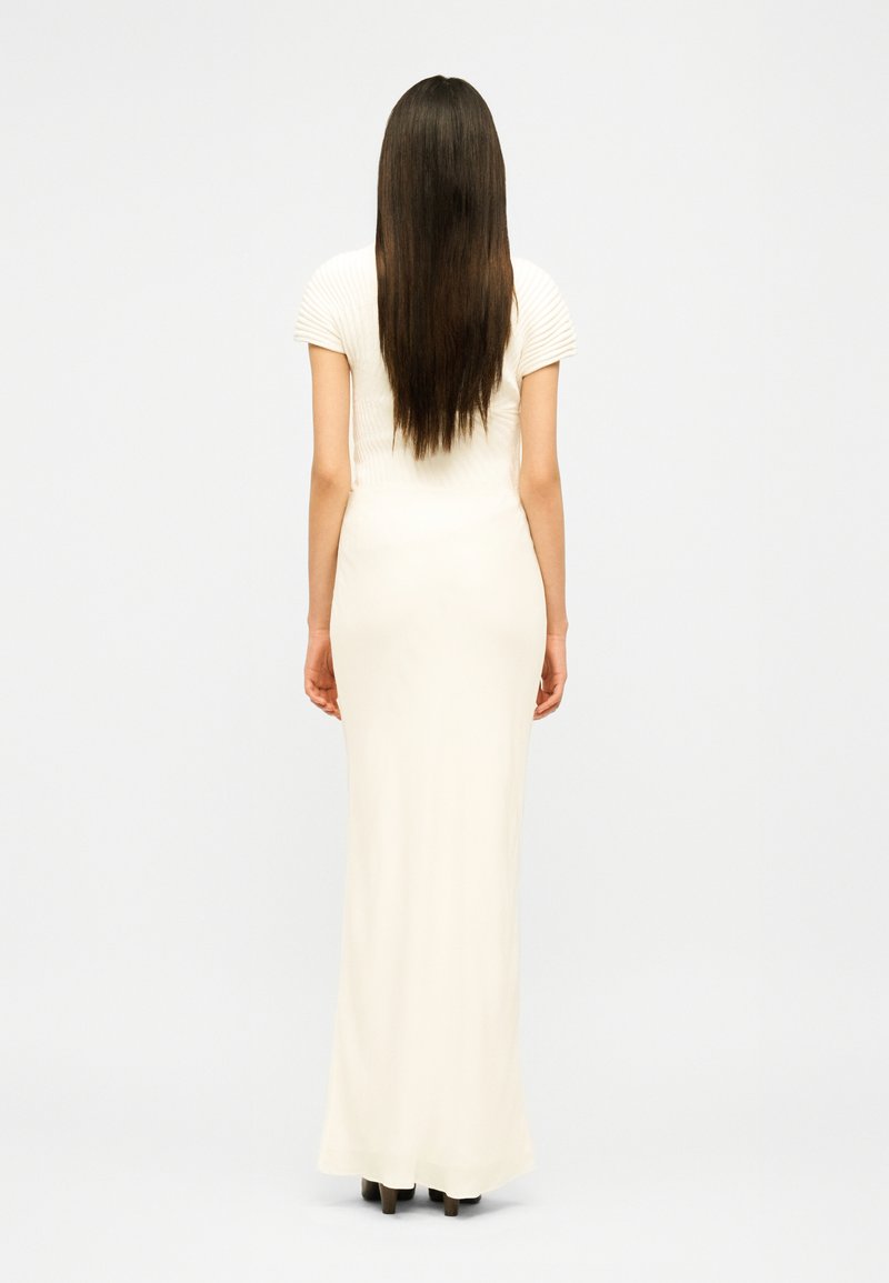 Femme aux longs cheveux bruns, vue de dos, portant une robe ivoire ajustée à manches courtes, longueur au sol, et des chaussures marron à talons.