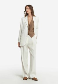 Vitt skräddarsytt jacka, brun väst med knappar, vita vida byxor och bruna sandaler med remmar. Slät tyg med en strukturerad design.