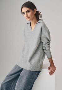 Maglione grigio lavorato a maglia con scollo a V e maniche arrotolate, abbinato a jeans larghi in denim scuro. Il tessuto sembra morbido con una leggera consistenza.