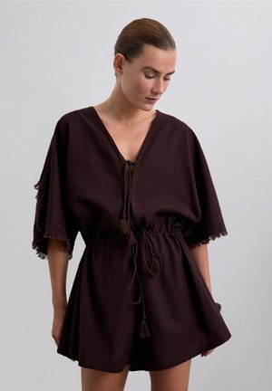 Mujer con el cabello peinado hacia atrás, vestida con un mono suelto de cuello en V marrón oscuro, con lazos de flecos y cintura fruncida, mirando hacia abajo contra un fondo liso.