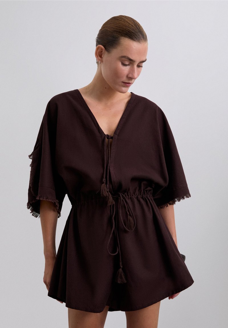 Vrouw met slicked-back haar, gekleed in een donkerbruin losse V-hals romper met franjes en een gatherings taille, kijkend naar beneden tegen een effen achtergrond.