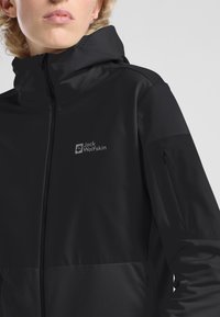 Giacca softshell nera con cappuccio, caratterizzata da un logo discreto sul petto, tasca con cerniera sulla manica e texture liscia.