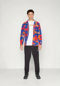 Fleecejakke i blå, rød og oransje geometriske mønstre med glidelås foran. Kombinert med svarte bukser og en hvit t-skjorte med tekst.