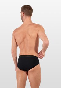 Schwarze Schwimmslips mit glatter Textur, elastischem Bund mit Branding und minimaler Bedeckung. Von hinten betrachtet.