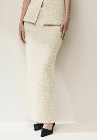 Robe tricotée texturée de couleur crème avec une silhouette ajustée, dotée d'une fente à la taille et accompagnée de chaussures noires à talons pointus.