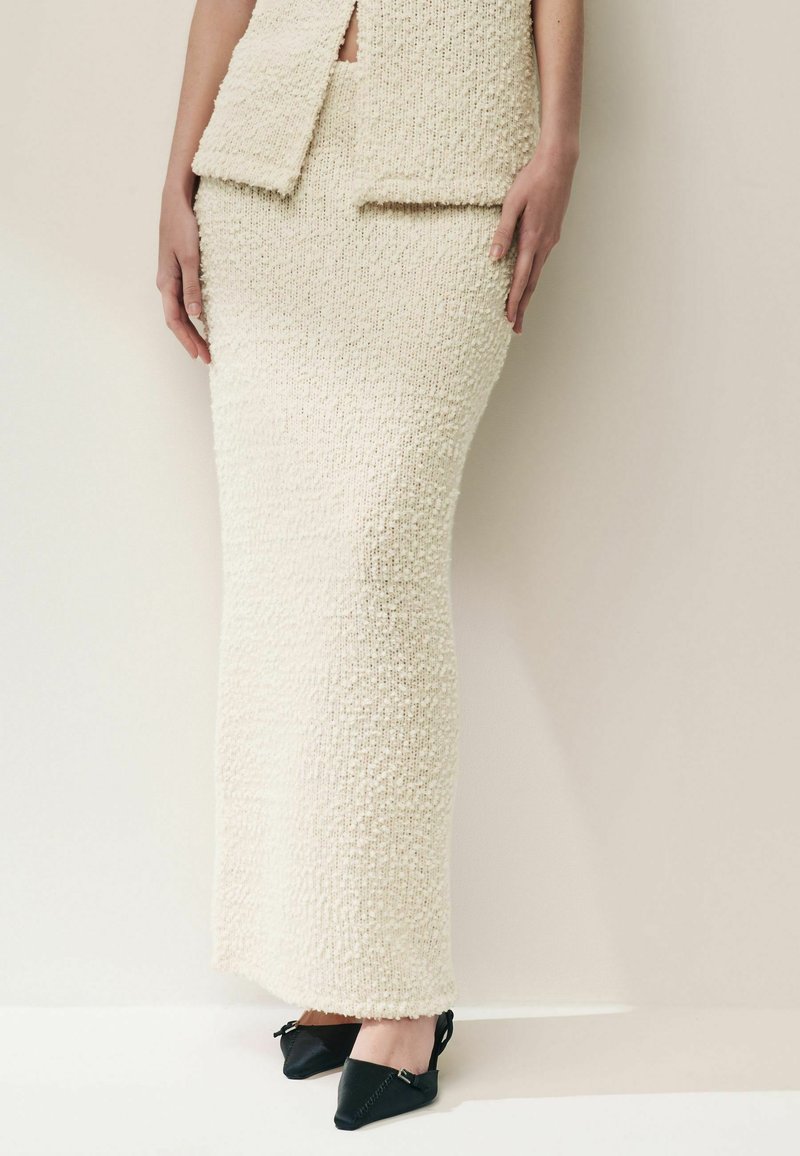 Robe tricotée texturée de couleur crème avec une silhouette ajustée, dotée d'une fente à la taille et accompagnée de chaussures noires à talons pointus.