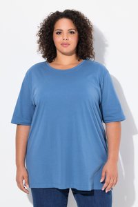 Donna con capelli ricci che indossa una maglietta blu larga e jeans scuri, in piedi davanti a uno sfondo bianco semplice.