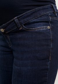 Mörkblå denimjeans med en åtsittande design, bältesöglor och en diskret knapp i kopparfärg med orange sömmar längs sömmarna.