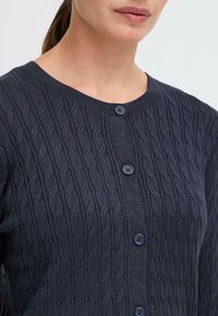 Mörkblå knappupp cardigan gjord av en texturerad stickad tyg med kabelmönster och ribbad halsringning. Har blå knappar.