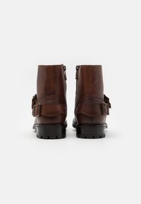 Botas de tobillo de cuero marrón con un acabado texturizado, que cuentan con una cremallera lateral, un hebilla ajustable y suelas negras resistentes.