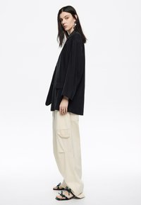 Blazer nero oversize con maniche lunghe, indossato sopra pantaloni cargo beige larghi. Sandali neri con accenti verdi e floreali completano l'outfit.