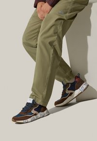 Le sneaker presentano pannelli in suede blu, marroni e grigi, una suola testurizzata e del testo bianco audace sul lato; indossate con pantaloni verde oliva.