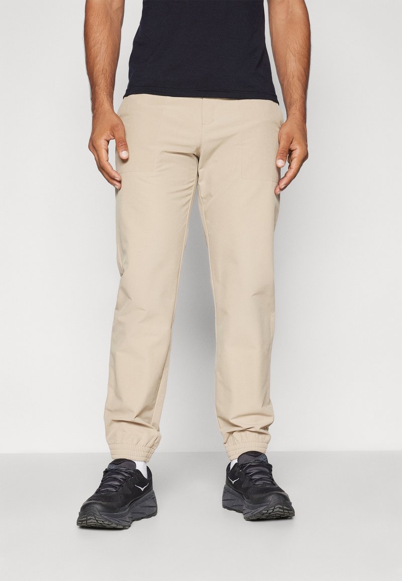 Béžové teplákové kalhoty vyrobené z měkkého materiálu, s elastickými manžetami a bočními kapsami, spárované s černými sportovními botami.