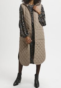 Mujer vestida con un chaleco largo acolchado beige sobre un vestido negro estampado, con botas negras hasta el tobillo, de pie frente a un fondo liso.