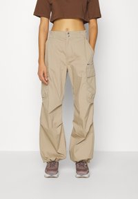 Pantalones cargo beige con un corte relajado, que presentan bolsillos laterales y dobladillo ajustado. La tela es ligera con una textura suave.
