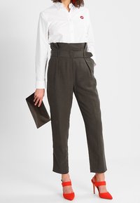 Kobieta w białej koszuli zapinanej na guziki i ciemnych spódnicach z wysokim stanem z paskiem, trzymająca czarną kopertówkę, w jasnoczerwonych szpilkach.