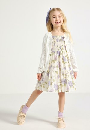 Hymyilevä nuori tyttö seisoo toisella jalalla edessä, pukeutuneena kukkakuosiseen mekkoon, valkoiseen neuleneuletakkiin, laventelinvärisiin sukkiin, beigenvärisiin kenkiin ja violettiin hiuskoristeeseen.