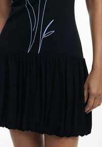 Rochie neagră cu un bust scurt și o fustă plisată, evazată. Prezintă broderie florală subtle în nuanțe de albastru deschis pe partea din față. Textură netedă.