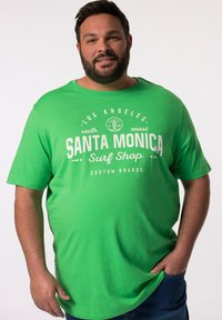Zelené tričko s krátkým rukávem a kulatým výstřihem, s bílým nápisem "Santa Monica Surf Shop" a grafikou. Měkká, lehká tkanina.