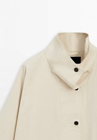 Beige jas met hoge kraag en zwarte knopen, getoond op een witte achtergrond, met focus op de bovenkant van de voorkant en de details van de kraag.