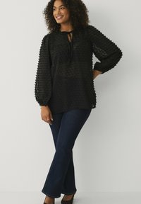 Zwarte lange mouwen blouse met een gestructureerd stippenpatroon, met een striknek, en licht opbollende mouwen, gecombineerd met donkere flared jeans en hakken.