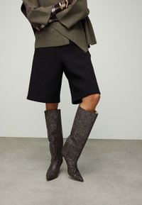 Bottes montantes argentées pailletées avec bouts pointus, surface texturée et ajustement serré, assorties à un short noir sur mesure et un haut vert olive.