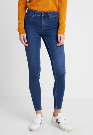 Osoba ubrana w niebieskie obcisłe dżinsy, białe trampki i pomarańczową bluzkę z długim rękawem stojąca na białym tle.