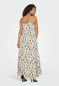 Maxi-jurk met dunne straps, witte stof met groene en zwarte geometrische patronen, vloeiende uitstraling, gecombineerd met zwarte hakken.