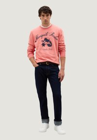 Sudadera rosa clara con el texto "Mesquite Rodeo" en azul marino, combinada con jeans de mezclilla azul oscuro y zapatillas blancas.