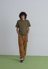 Tricou scurt de culoare verde măsliniu, pantaloni largi maro și adidași negri cu accente albe, stând pe un podea verde deschis.