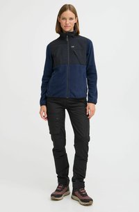 Veste en polaire avec un panneau supérieur noir et un bas bleu marine, fermeture éclair intégrale, col montant, assortie à un pantalon cargo noir et des bottes de randonnée.