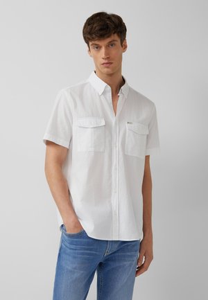 Jeune homme aux cheveux courts et bruns portant une chemise blanche à manches courtes avec poches, et un jean bleu, debout avec une main dans la poche.