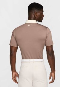 Brūns īsajām piedurknēm polo krekls ar krēmkrāsas apkakli un Nike logo uz aizmugures. Nēsāts ar gaišu biksēm un baltu jostas. Gluda tekstūra.