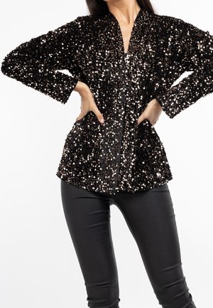 Kobieta w czarnej cekinowej kurtce i czarnych skórzanych spodniach, stojąca z rękami na biodrach na białym tle.