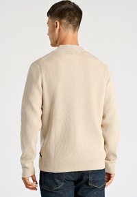 Beige trui van geribbelde stof met lange mouwen en een ronde hals, met een licht verlengde schoudernaad en een contrasterend logolabel aan de zijkant.