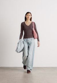 Top lung cu mâneci lungi de culoare maro, blugi largi decolorate ușor în nuanță albastră cu detalii pe lateral, geantă roz și pantofi maronii. Fundal simplu, model mergând înainte.