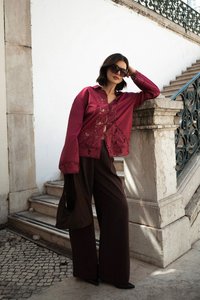 Burgunderrot bestickte Bluse mit langen Ärmeln, kombiniert mit weiten braunen Hosen. Das Model lehnt an einer Steinmauer und trägt dunkle Sonnenbrillen.