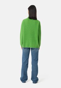 Grüner übergroßer Strickpullover mit hohem Kragen, ausgestattet mit Rippstruktur, kombiniert mit hellblauen Schlagjeans. Rückenansicht, minimaler Hintergrund.
