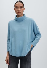 Eine Frau trägt einen hellblauen, lockeren Rollkragenpullover und dunkelgraue Jeans und steht vor einem schlichten hellgrauen Hintergrund.