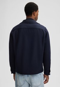 Veste en polaire bleu marine avec un col classique, dotée de poignets boutonnés et d'un design épuré. Couture visible sur les épaules, texture douce.