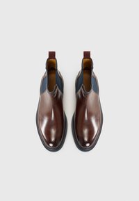 Des chaussures brunes en cuir à enfiler présentent une finition brillante, des accents bleu marine sur les côtés et une tirette bordeaux au talon. Semelle en caoutchouc pour une bonne adhérence.