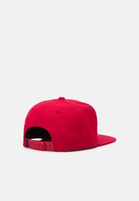 Sarkans snapback cepure ar vienkāršu dizainu, teksturētu audumu, apaļu augšu un regulējamu siksniņu ar vairākiem izmēru caurumiem aizmugurē.